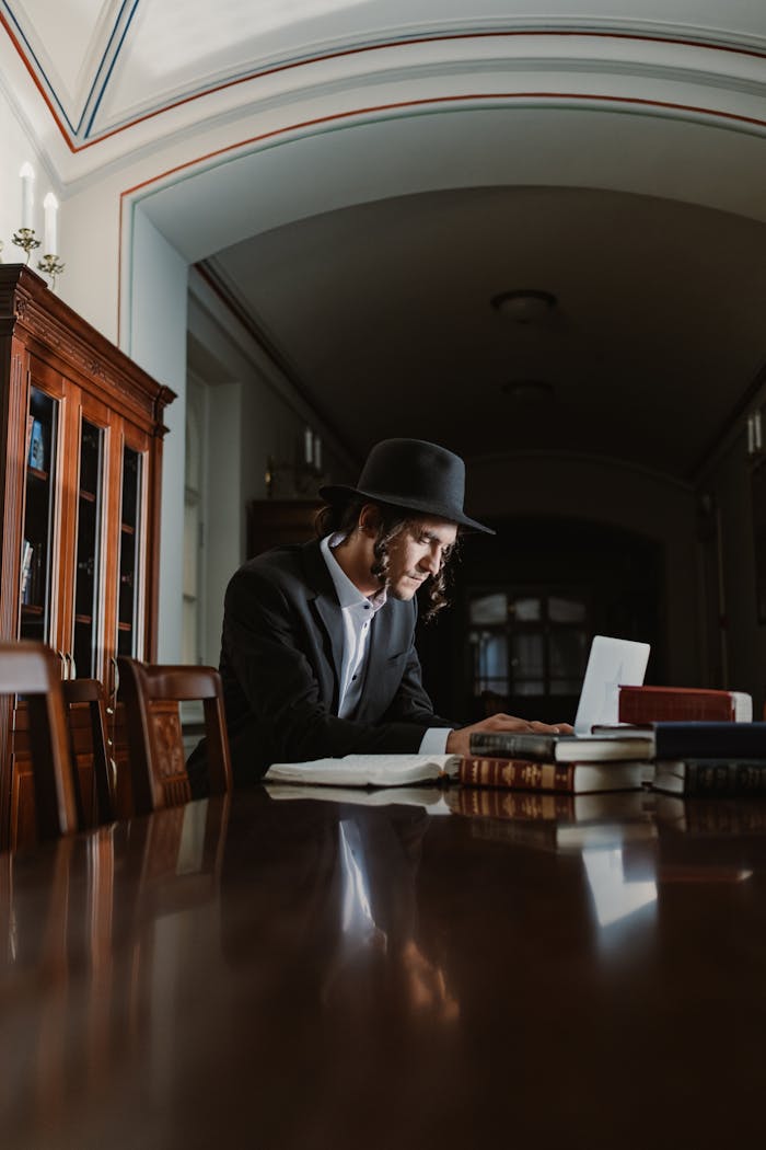 story-02 Orthodox Jewish man with hat and payot studying religious texts indoors on a wooden table.